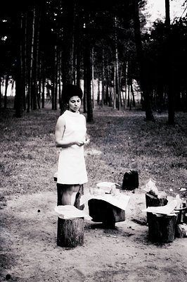 A young woman in a short, sleeveless dress stands amidst a pine forest setting. A makeshift table of tree stumps and planks d...