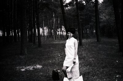 A woman in a short, A-line dress and elaborate beehive hairstyle smiles while walking through a dense pine forest. She carrie...
