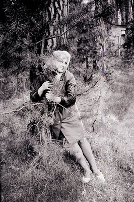 A young woman sits amidst tall grasses, partially obscured by pine branches. She wears a short-sleeved dress and stockings, c...