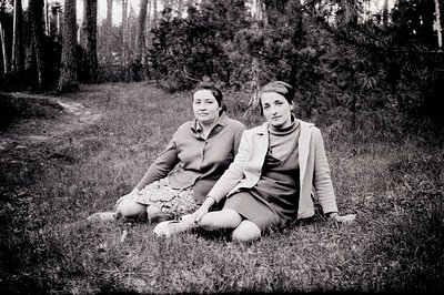 Two young women seated in tall grass, framed by a pine forest. Both wear 1960s-era clothing: a patterned shirt and skirt, and...