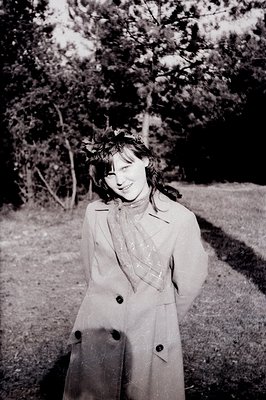 A young woman wearing a double-breasted trench coat and a floral crown smiles directly at the camera. Likely 1960s or 70s, th...