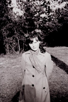 A young woman stands in a wooded area, wearing a double-breasted trench coat and patterned scarf. Floral accents adorn her ha...