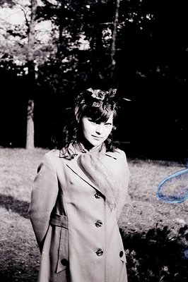B&W portrait of a young woman wearing a double-breasted coat and floral headband. A sheer scarf is knotted at the neck. Likel...