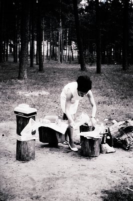 A woman in a short, white dress and heels appears to be preparing a picnic amongst tree stumps in a wooded area. Vintage cool...