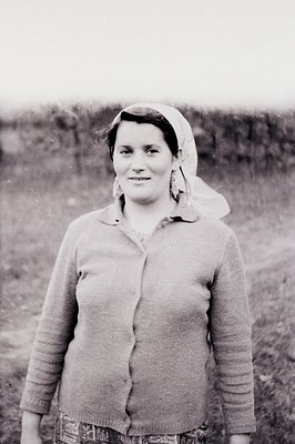 Portrait of a woman wearing a hand-knitted cardigan, patterned skirt, and headscarf. Traditional style suggesting a rural set...