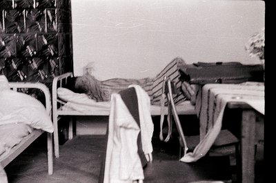 A stark, institutional dormitory space features rows of metal bunk beds. Visible linens and bedding suggest a period of disus...