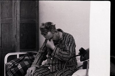 Man sits on a bed, appearing distressed, in striped pajamas. A vintage, adjustable desk lamp is visible on a side table. Star...