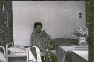 A young woman with a bob haircut sits bundled in a patterned robe, seemingly lost in thought. A bedside table holds a vase of...