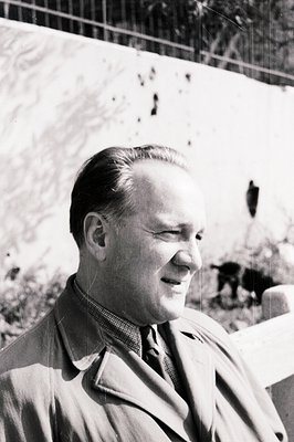 A man in a trench coat and tie smiles slightly in a profile view. The image, likely a candid portrait, showcases classic mid-...