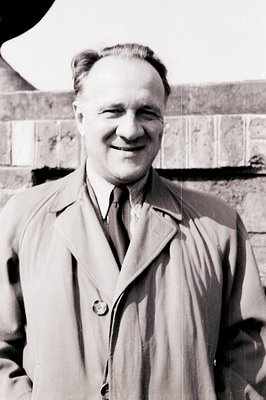 Portrait of a gentleman in a trench coat and tie, smiling directly at the camera. Likely taken outdoors with a textured brick...