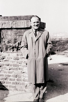 Man in a trench coat stands against a weathered brick wall with graffiti, a subtle smile on his face. Appears to be a mid-cen...