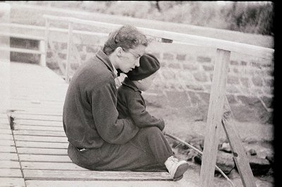A young man sits pensively on a weathered wooden structure, possibly a dock or bridge. He wears a dark jacket and a hooded sw...