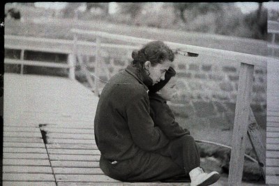 A seated individual with short hair gazes downward, appearing contemplative, on a wooden structure overlooking water. The arc...
