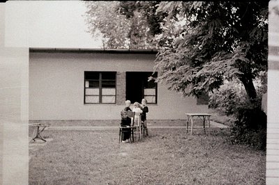 A group of children, possibly students, are gathered outdoors in front of a simple, modernist building with large windows. Th...