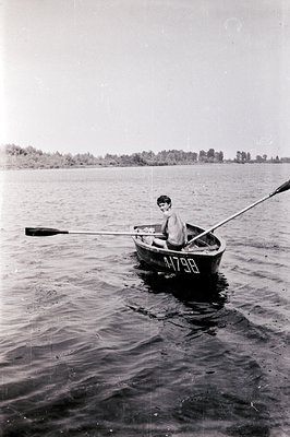 A young boy rows a small, wooden boat across a body of water. The boat’s hull is marked with "4-1798." Treeline visible in th...