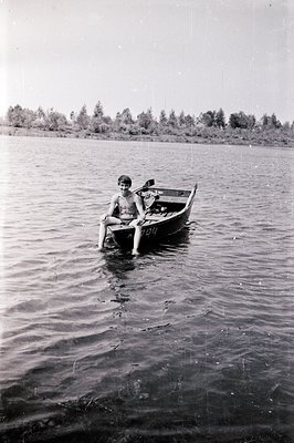 Two young boys seated in a small rowboat afloat on a calm lake, surrounded by lush greenery. The image, likely a snapshot, ex...