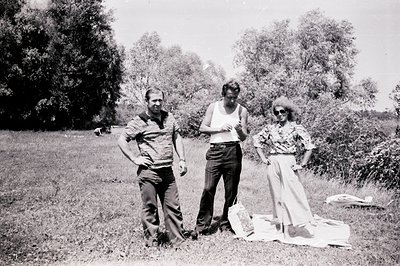 Three figures stand in a grassy, tree-lined field. The man at left wears a patterned polo shirt, the center figure a white ta...