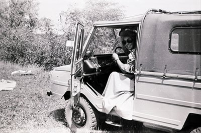 Young woman in a long dress and sunglasses is driving an early model military-style vehicle, likely a UAZ-69. Tall grasses an...