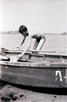 A young boy in dark swim trunks leans over the bow of a small wooden rowboat on calm water. The boat’s hull shows signs of we...