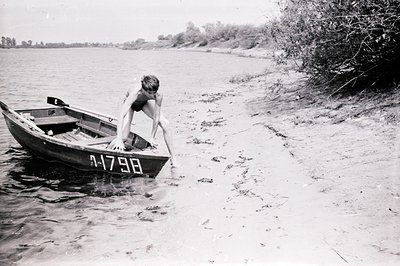 A young man in swimming trunks emerges from a small rowboat near a riverbank. The boat, marked "1-1798", rests partially subm...