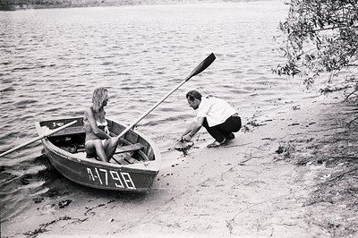 A young couple, likely in the 1960s, occupies a small rowboat pulled ashore. The woman wears a modest swimsuit; the man is fo...