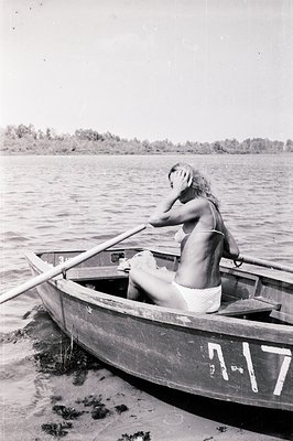 Woman in a small rowboat, seen from behind, holds an oar. Boat number "7-17" is visible on the hull. Lakeside setting with tr...