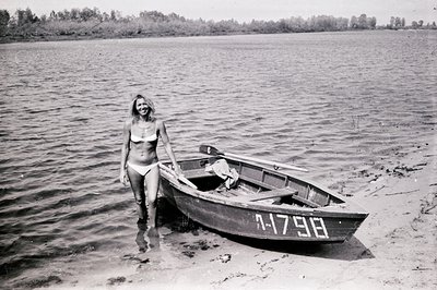 A young woman in a white bikini stands ankle-deep in water beside a small rowboat with the number "1-17-98" painted on its si...