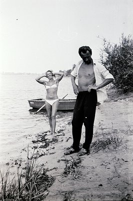 A man and woman stand by a small rowboat on a sandy shoreline. He’s partially undressed, revealing his torso, and she wears a...