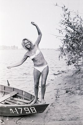 A young woman in a white bikini stands on a small rowboat, arms raised in a jubilant pose. Sandy beach and calm water backdro...