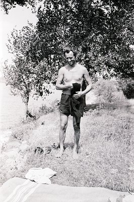 A shirtless man stands on a grassy hillside, partially obscured by a leafy tree. He holds a dark, draped cloth around his wai...