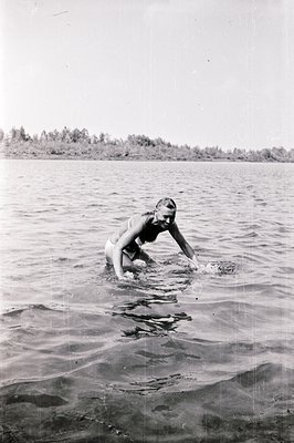 A young woman swims in a calm body of water, appearing to dog paddle. The shoreline and treeline are visible in the distant b...