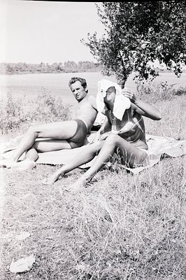 A monochrome portrait captures a man and woman, both nude, reclining on a blanket amidst tall grass and foliage, likely near ...