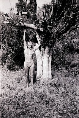A woman hangs from a tree branch, smiling directly at the camera. Set in a field of tall grass and shrubbery, the image featu...