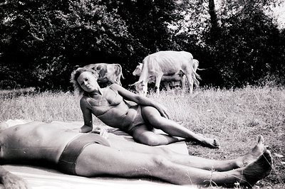 A striking black and white image depicting two figures reclining on a white sheet amidst tall grasses and foliage. One figure...