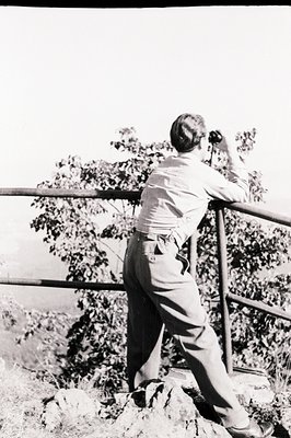 Man leans against a metal railing overlooking a landscape, using binoculars. He wears a light-colored shirt and trousers. Roc...