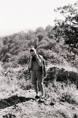 Man in shorts and a jacket, sporting a camera around his neck, hikes up a slope overgrown with foliage. The scenery suggests ...