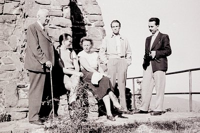 Group portrait of five individuals near a stone wall, possibly a fortification or ruin. A mature man with a cane stands with ...