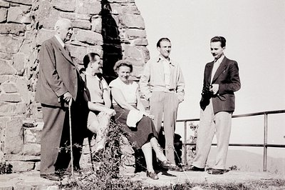 A group of five individuals poses outdoors, near a stone structure possibly a wall or retaining wall. Four are formally dress...