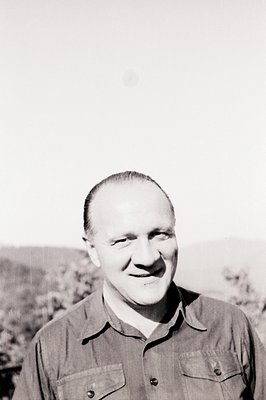 Close-up portrait of a man smiling directly at the camera. He wears a collared shirt with pocket flaps. The background featur...