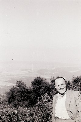 A smiling man in a tweed jacket stands amidst lush greenery, overlooking a hazy landscape. The photo exhibits characteristic ...