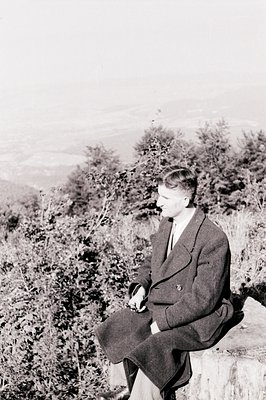 A seated man in a dark overcoat and tie poses on a stone ledge with a blurred, forested landscape behind. Likely a portrait f...