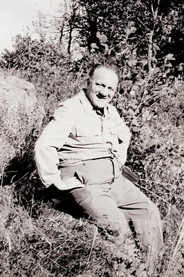 A man with a jovial smile sits casually on a rocky outcrop in a grassy field. He's dressed in a collared shirt and slacks, ap...