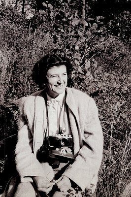 A smiling woman, likely a photographer, sits amidst tall grass and foliage. She wears a suit jacket & tie, with a vintage cam...
