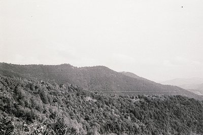 Dense, forested hills cascade across the frame, receding into a hazy distance. The landscape shows signs of terracing, sugges...