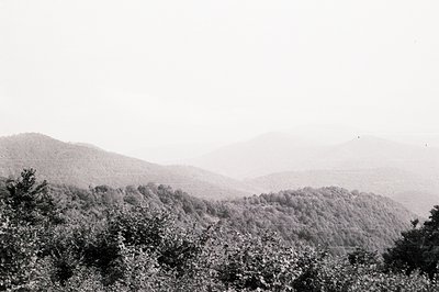 Dramatic, layered mountain range receding into atmospheric haze. Dense, forested slopes fill the foreground, contrasting shar...