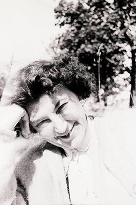 A portrait of a smiling woman with a curled hairstyle, likely 1950s-1970s. She wears a collared shirt and rests her hand on h...