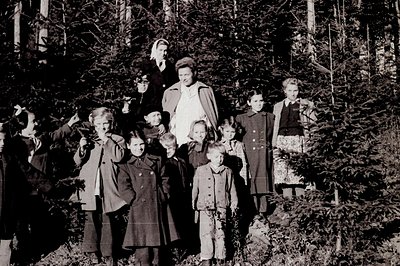 A group portrait of 13 children and two adults, posed outdoors among evergreen trees. The children are formally dressed in co...