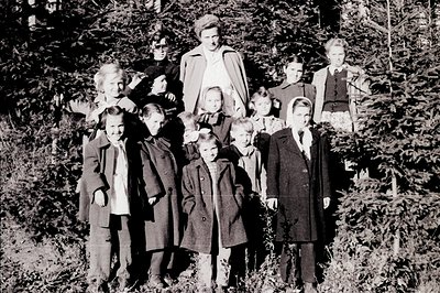 A group of children and two adults pose amongst evergreen trees. The adults, a man in an overcoat and a woman with a headscar...