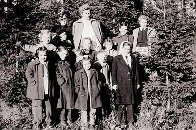 Group portrait of children and a young woman, likely outdoors near a forest. Subjects wear 1960s-70s outerwear. The woman is ...