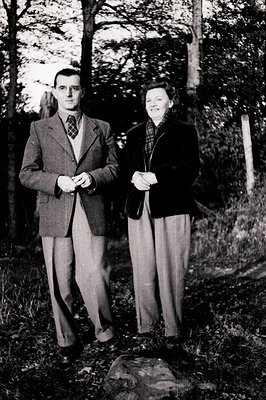 A formally dressed man and woman stand outdoors amidst foliage. Both wear suits and ties/scarves, suggesting a special occasi...